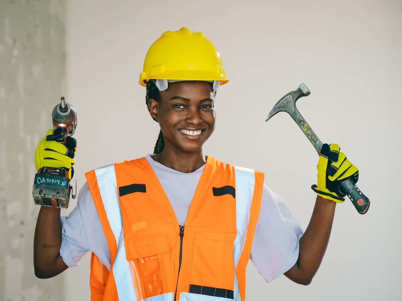 Smiling female engineer wearing safety gear, holding a drill and hammer confidently.